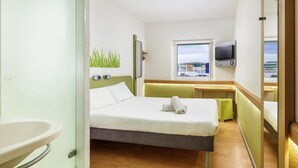 A hotel room with a bed, a bedside table, a TV mounted on the wall, and a window with a view of buildings. - ibis budget Bergerac (Bergerac)