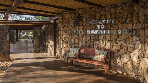 Een stenen muur met een bord dat Safari Lodge verwelkomt, een houten bankje en een betonnen pad dat verder leidt.