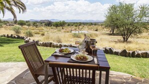 Een buitentafel met borden eten, een karaf en glazen, met op de achtergrond een grasveld en bomen.