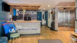 A reception area with a counter, a blue couch, a small table, a wall clock, and an elevator. - ibis budget Meudon Paris Ouest (Meudon)