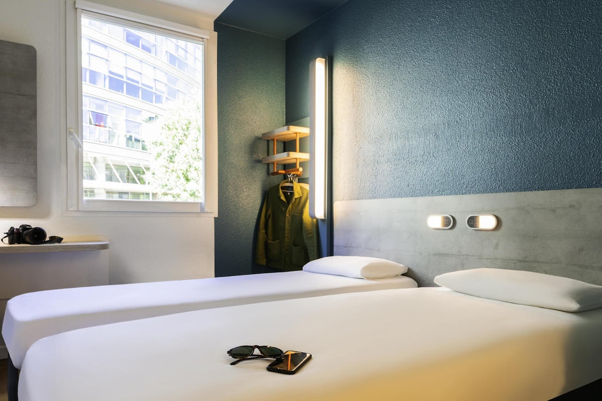 A hotel room with two beds, a window with a view of buildings, a coat rack with a jacket, and a bedside table with a camera.