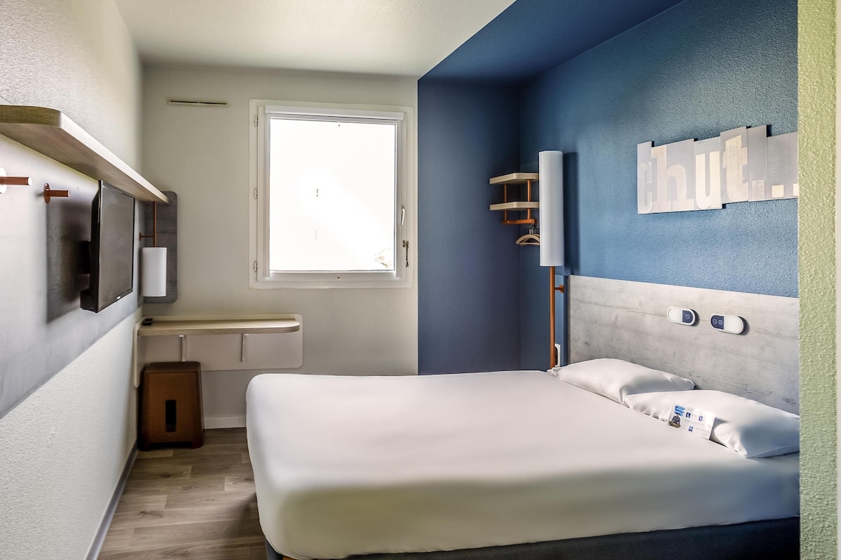 A modern hotel room with a bed, bedside table, window, and a wall-mounted light fixture.