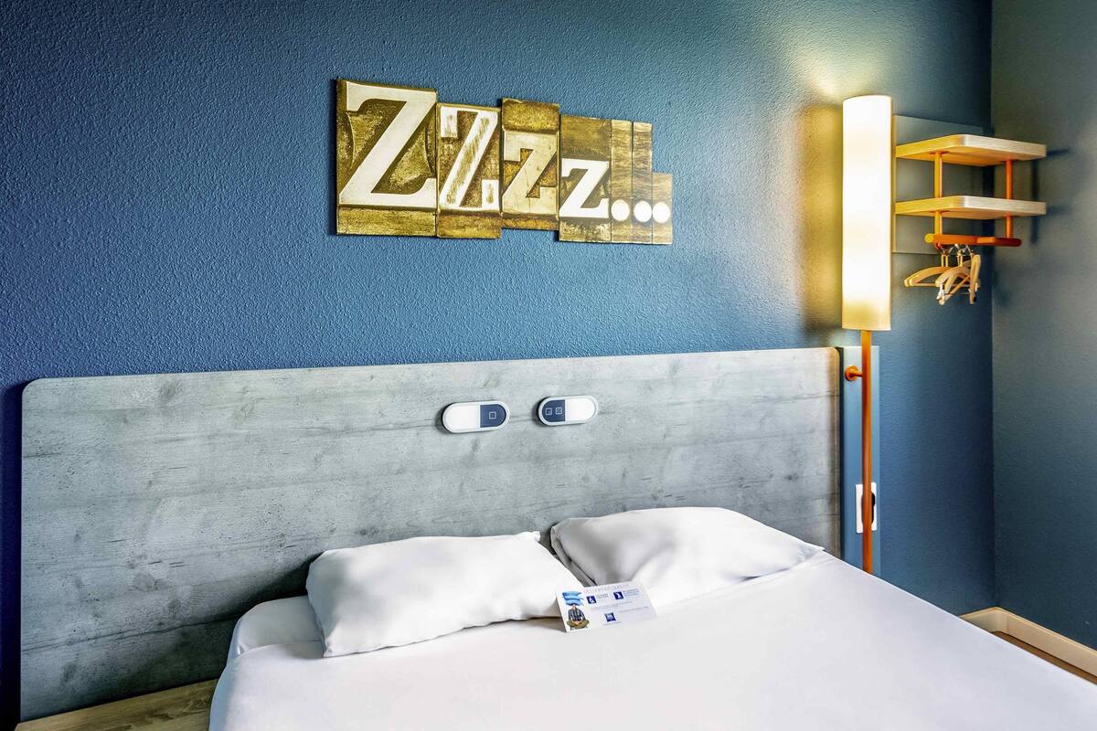 A hotel room with a bed, a headboard, a wall-mounted light, and a decorative wall piece.