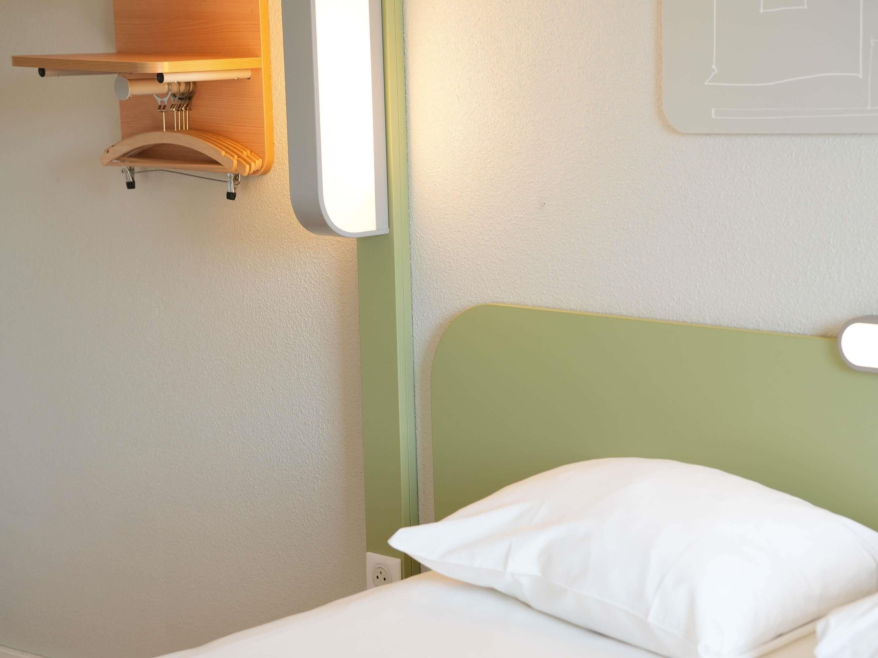 En enkeltseng med et grønt sengegavl, en træhylde over og en vægmonteret lampe.