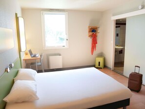 A hotel room with a bed, a desk with a laptop, a radiator, a small green cabinet, a suitcase, and a bathroom visible through an open door. - ibis budget Caen Porte de Bretagne (Bretteville-sur-Odon)