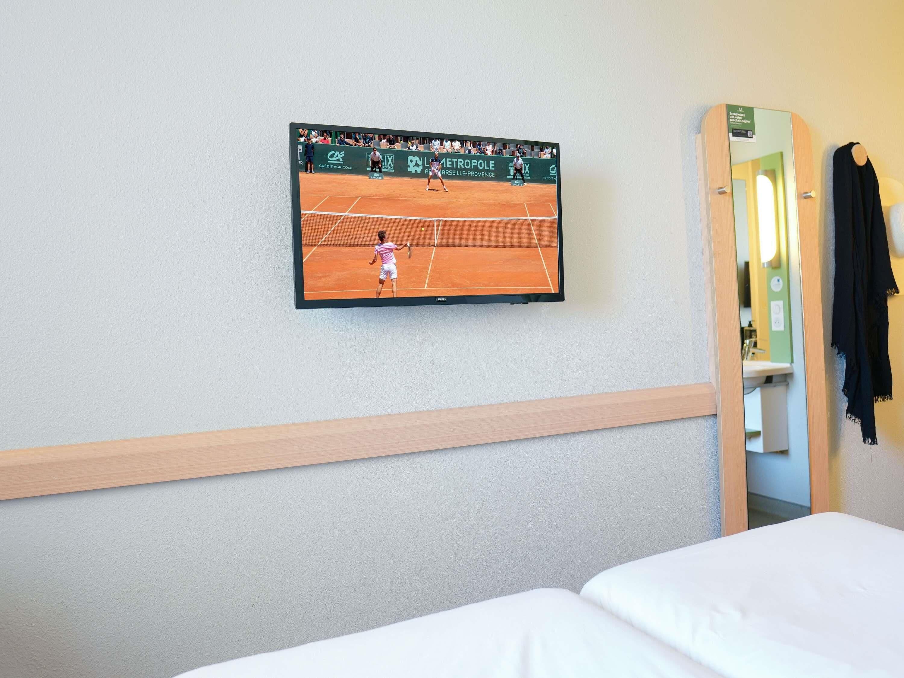 A hotel room with a television displaying a tennis match, a bed, and a mirror.
