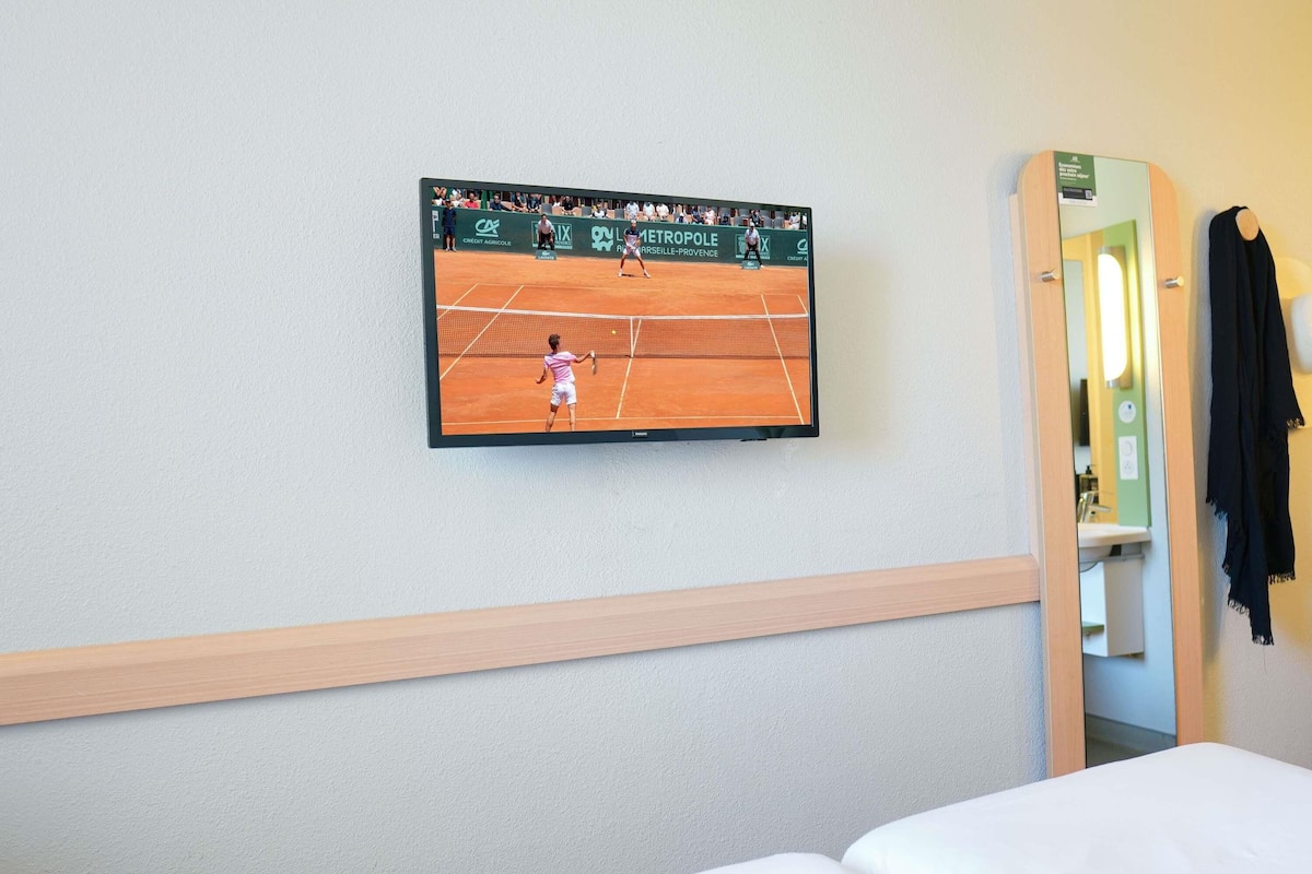 Ein Hotelzimmer mit Fernseher, der ein Tennismatch zeigt, einem Bett und einem Spiegel.