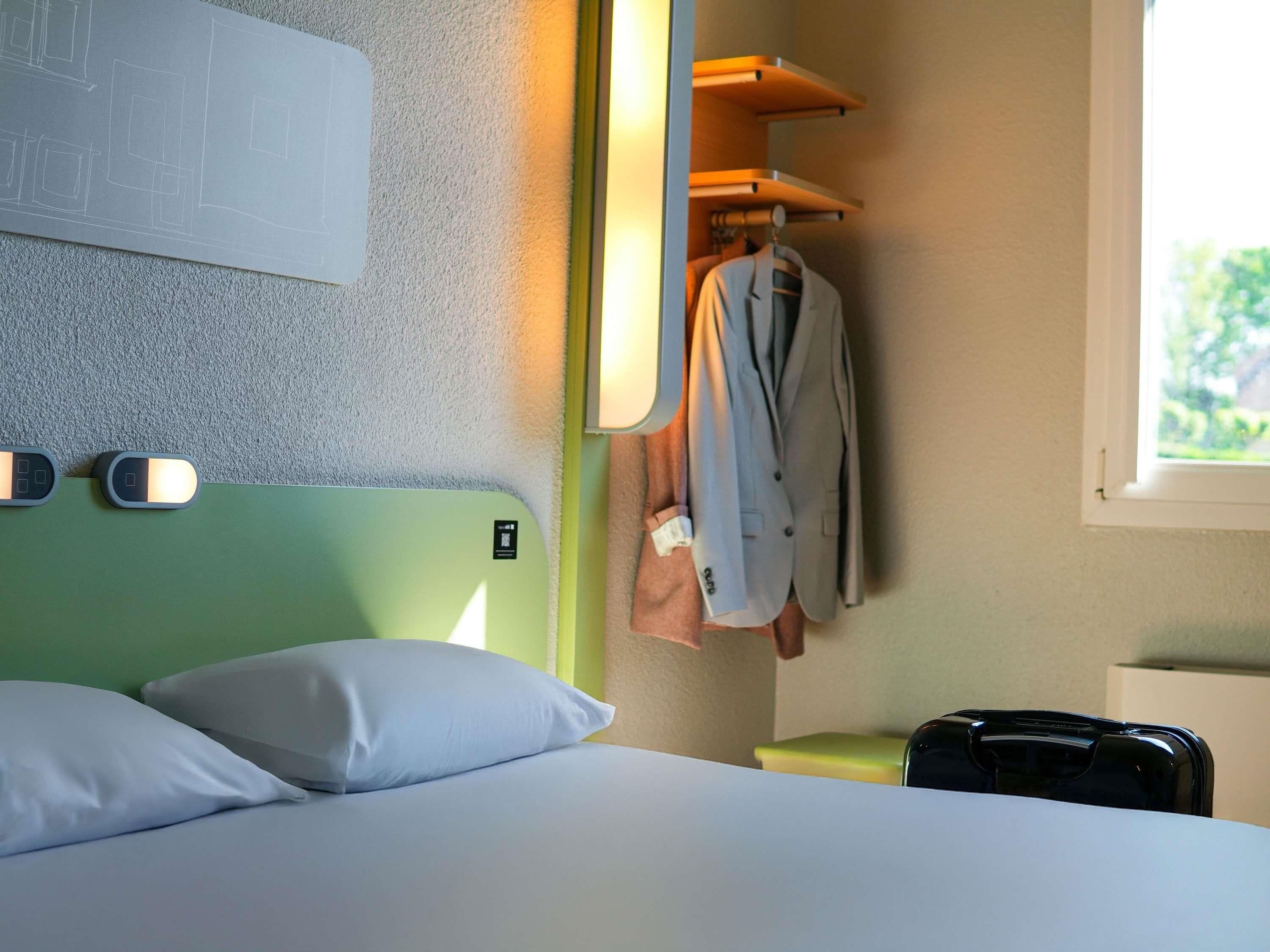 A hotel room with a green headboard, a bed with white pillows, a hanging coat rack with a suit, a suitcase, and a window with a view of greenery.