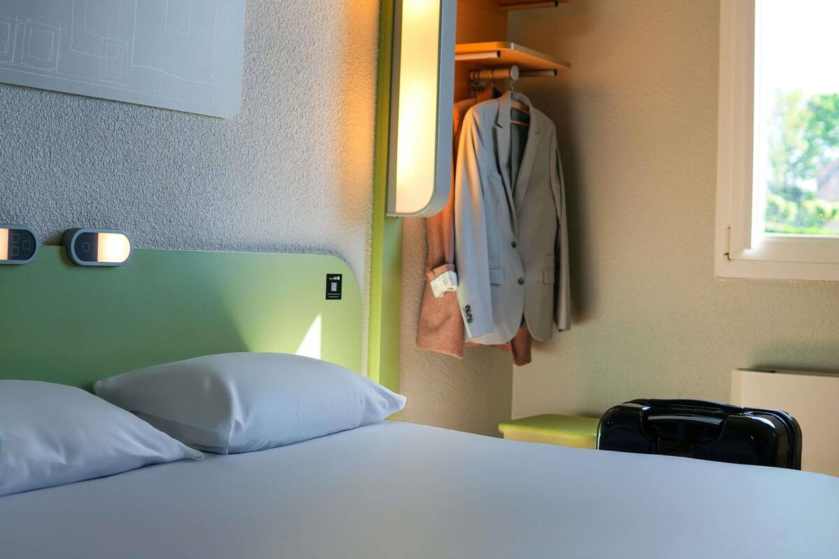 Ein Hotelzimmer mit einem grünen Kopfteil, einem Bett mit weißen Kissen, einem Kleiderständer mit einem Anzug, einem Koffer und einem Fenster mit Blick auf Grünflächen.