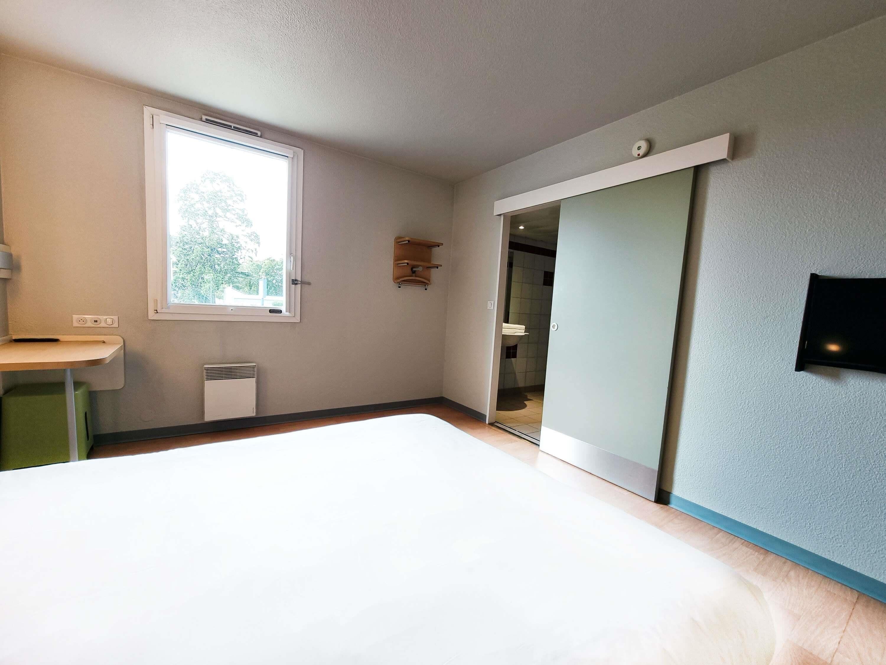 Une chambre à coucher comprenant un lit, une fenêtre, une table de chevet, un téléviseur fixé au mur et une porte donnant sur une salle de bain.