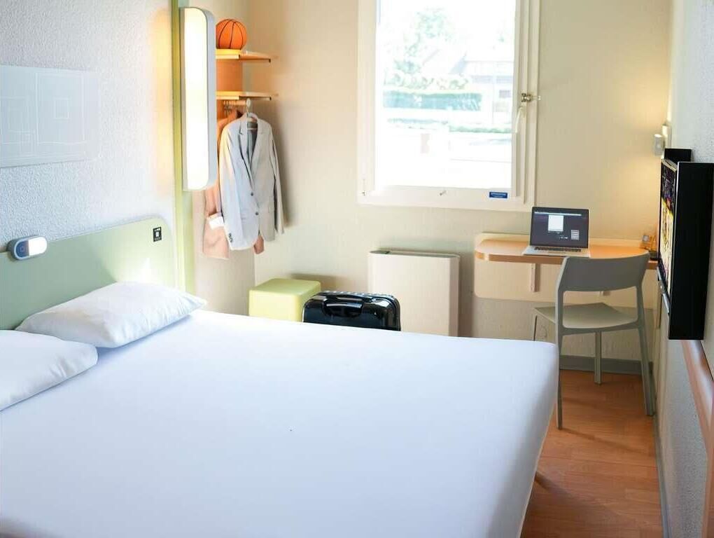 Une chambre d’hôtel avec un lit, un bureau avec un ordinateur portable, une chaise, un radiateur et une fenêtre.