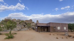Un bâtiment moderne doté d’un toit plat et d’une entrée abritée, entouré de gros rochers et d’une végétation clairsemée.