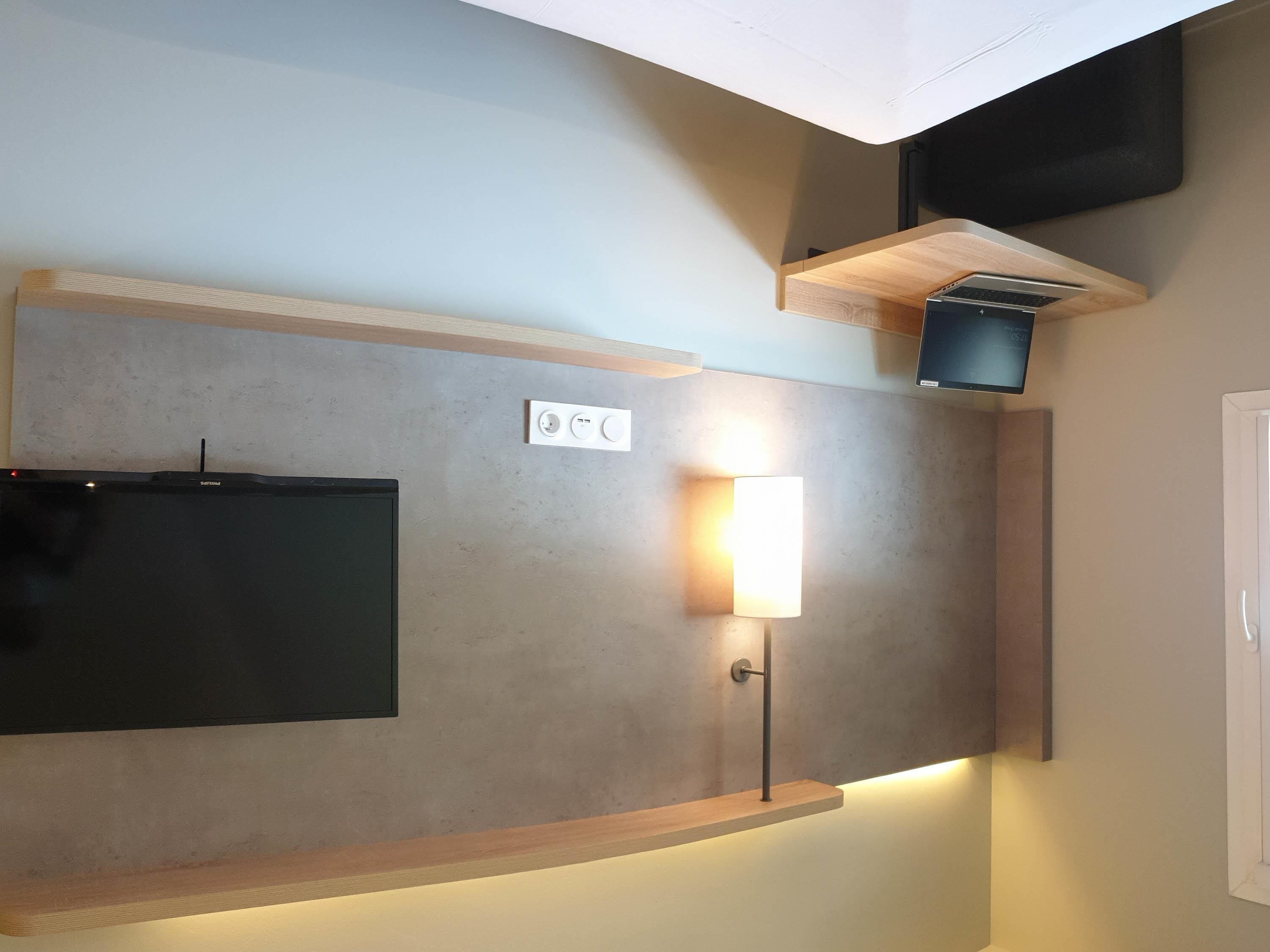 A modern room with a flat-screen TV mounted on the wall, a wall-mounted light fixture, and a wooden shelf above the TV.