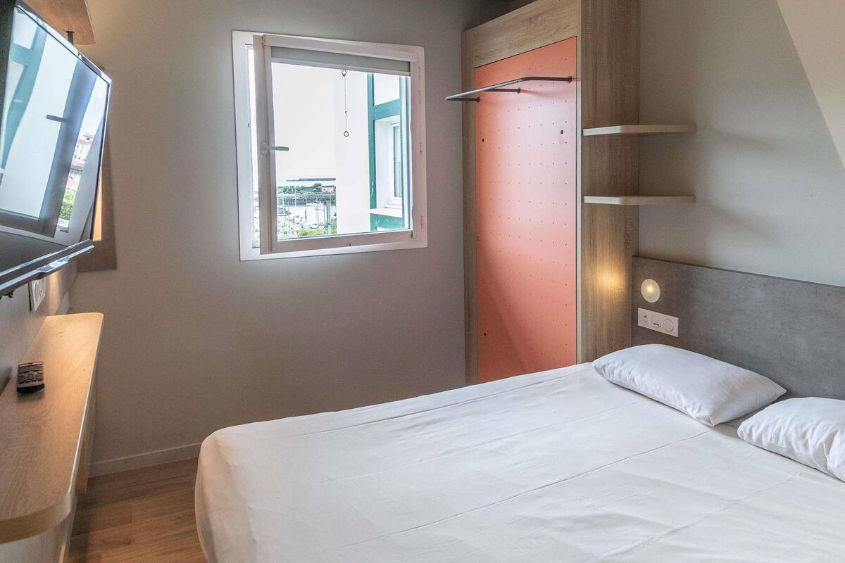 A bedroom with a bed, a TV on a stand, a window, and a door with a towel rack.