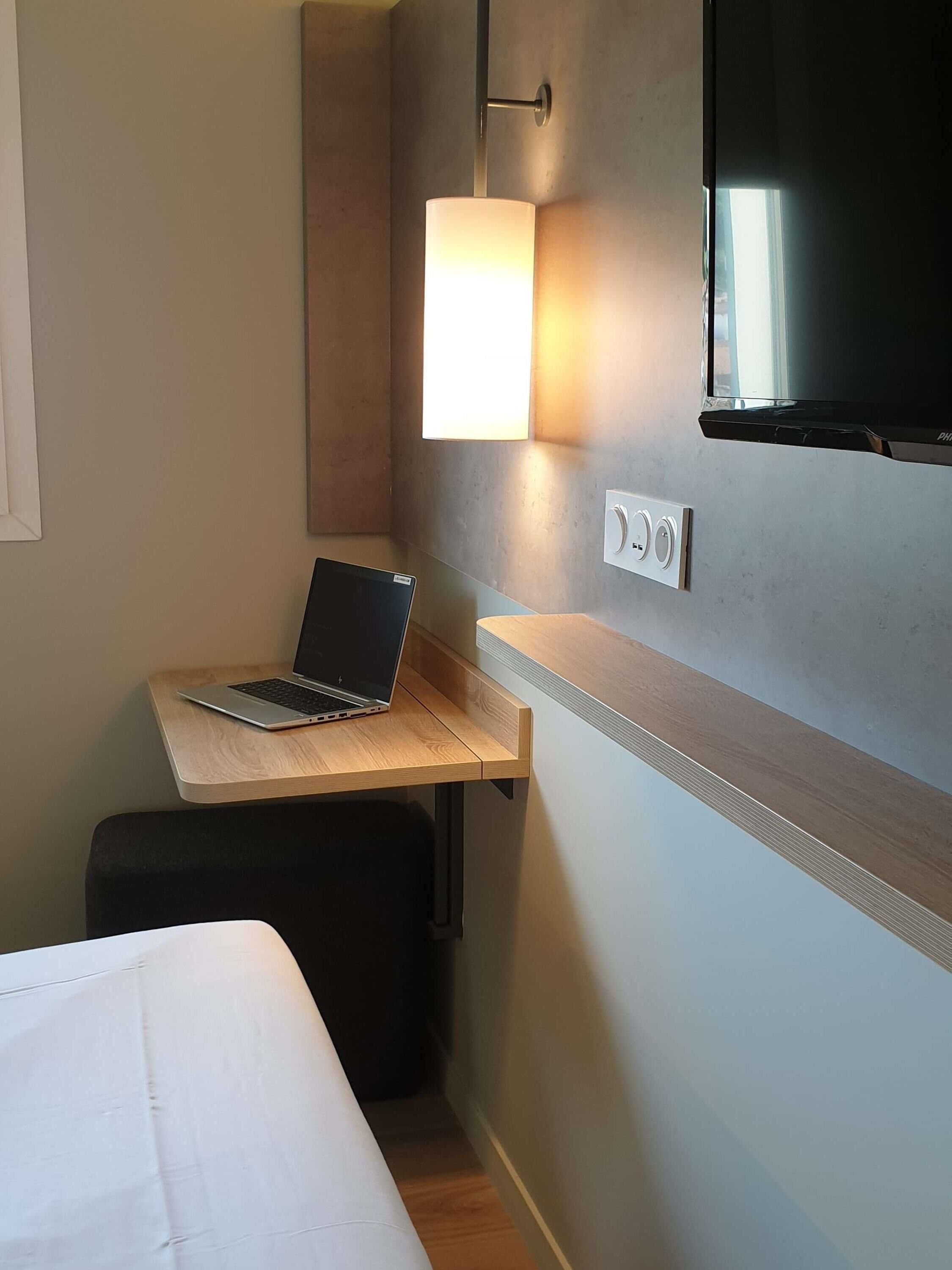 Una habitación de hotel con cama, un escritorio con una computadora portátil, un televisor montado en la pared y una mesita de noche.