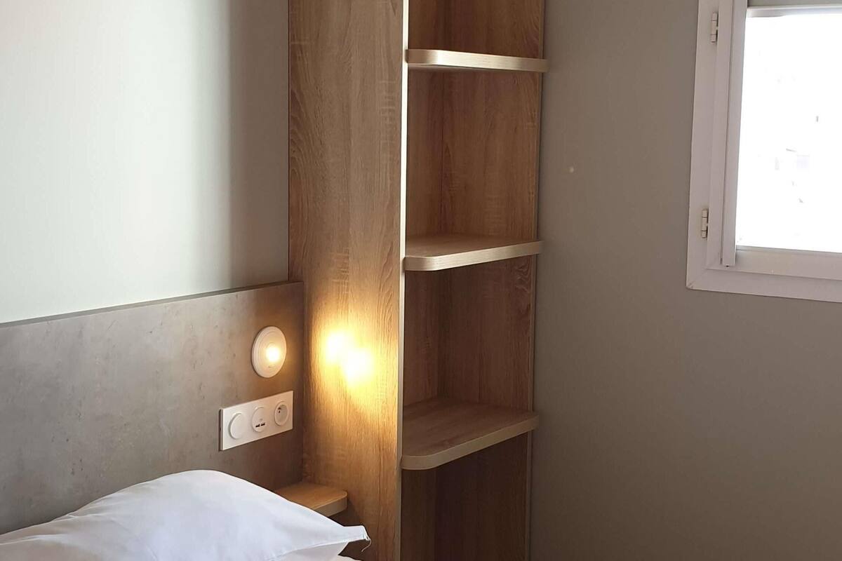 A bedroom with a bed, a wooden shelf unit, a wall socket, and a window.