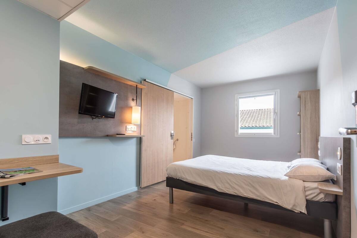 A bedroom with a bed, a desk, a TV mounted on the wall, and a window with a view.