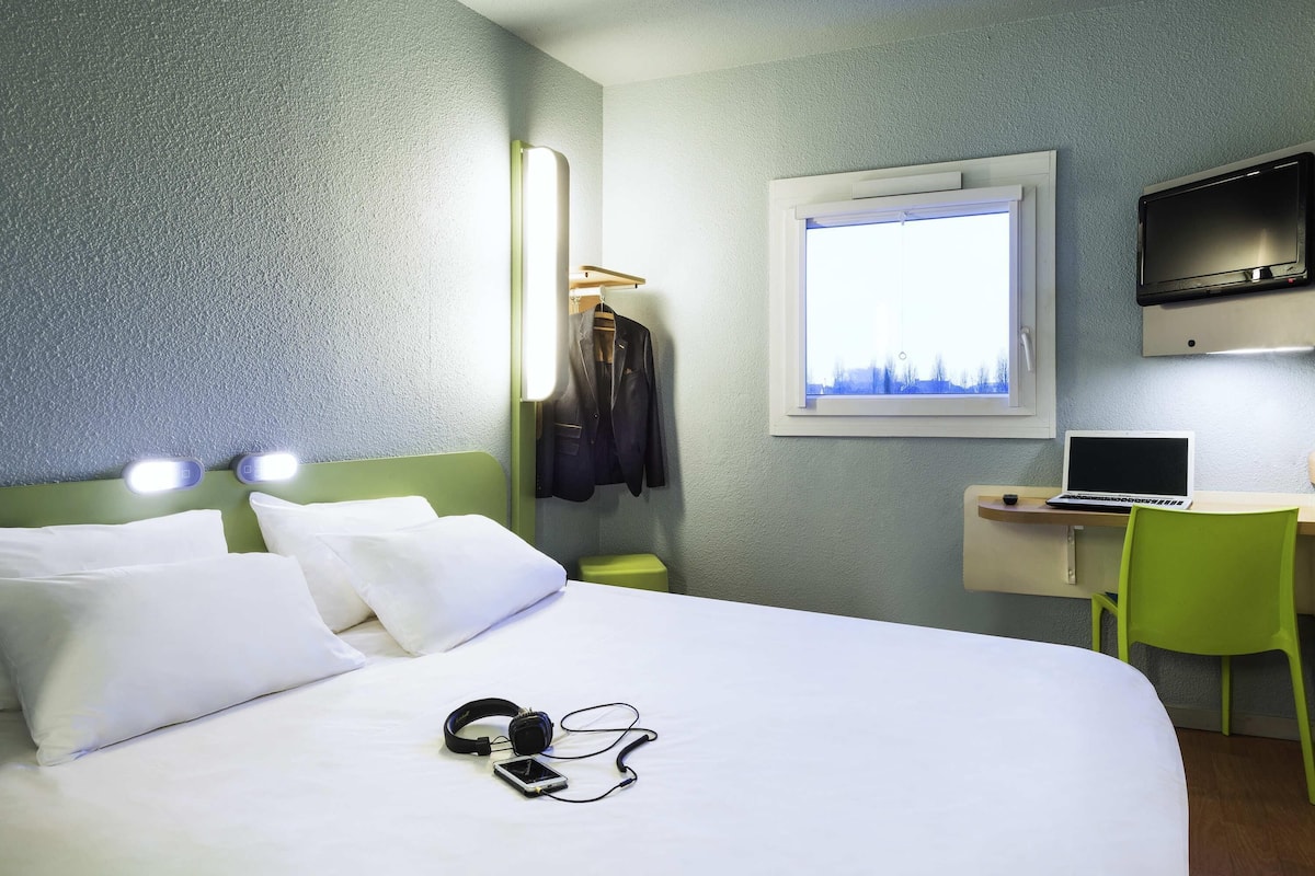 Ein Hotelzimmer mit Bett, Schreibtisch mit Laptop, Fernseher, Fenster mit Aussicht und Kleiderständer mit Jacke.
