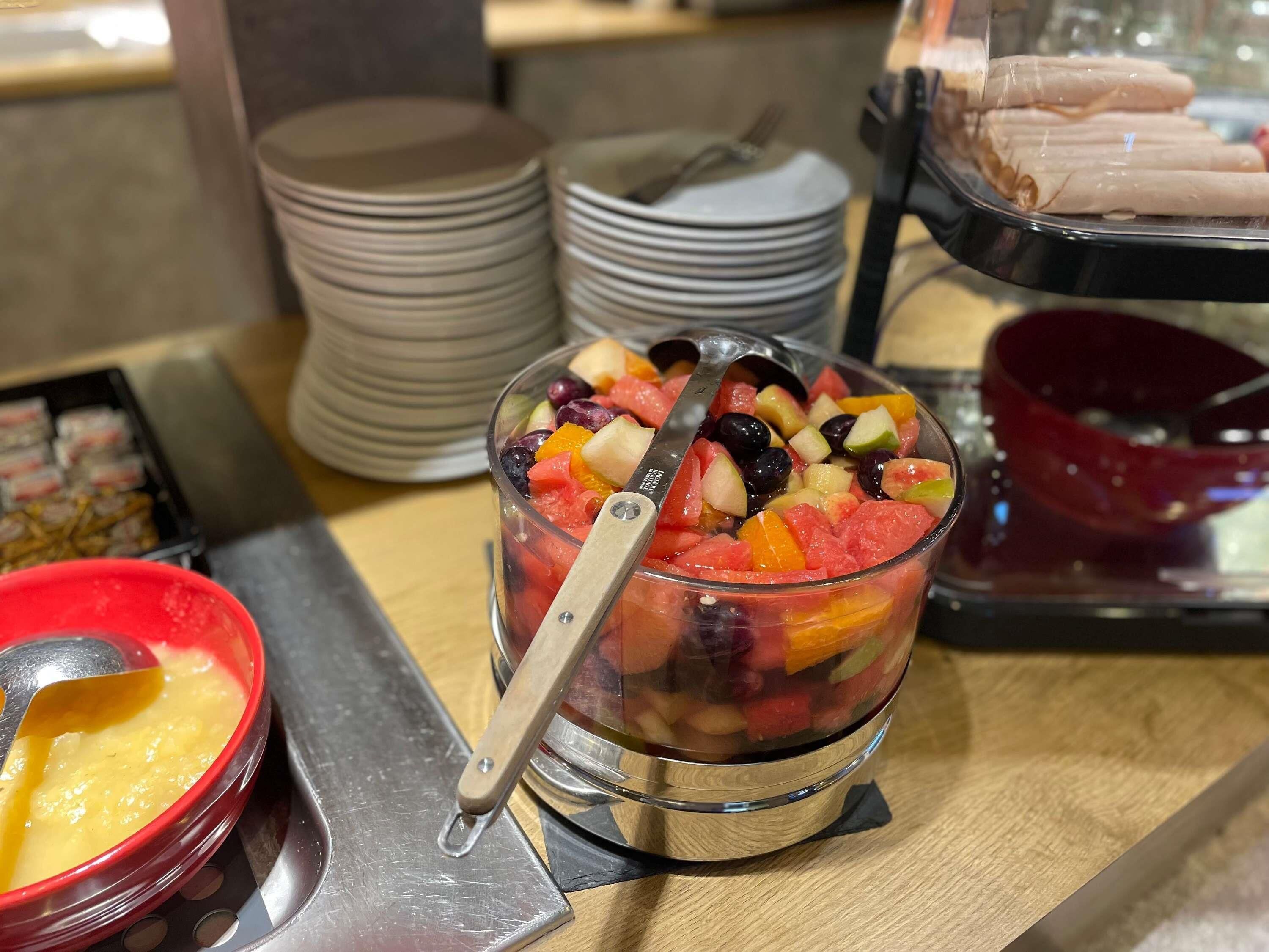 A buffet with a fruit bowl containing various fruits and a spoon, surrounded by stacks of plates and bowls.