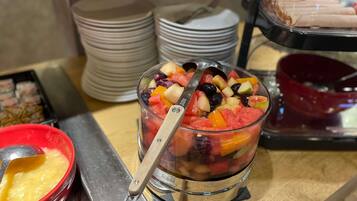 Een buffet met een fruitschaal vol verschillende soorten fruit en een lepel, omringd door stapels borden en kommen.