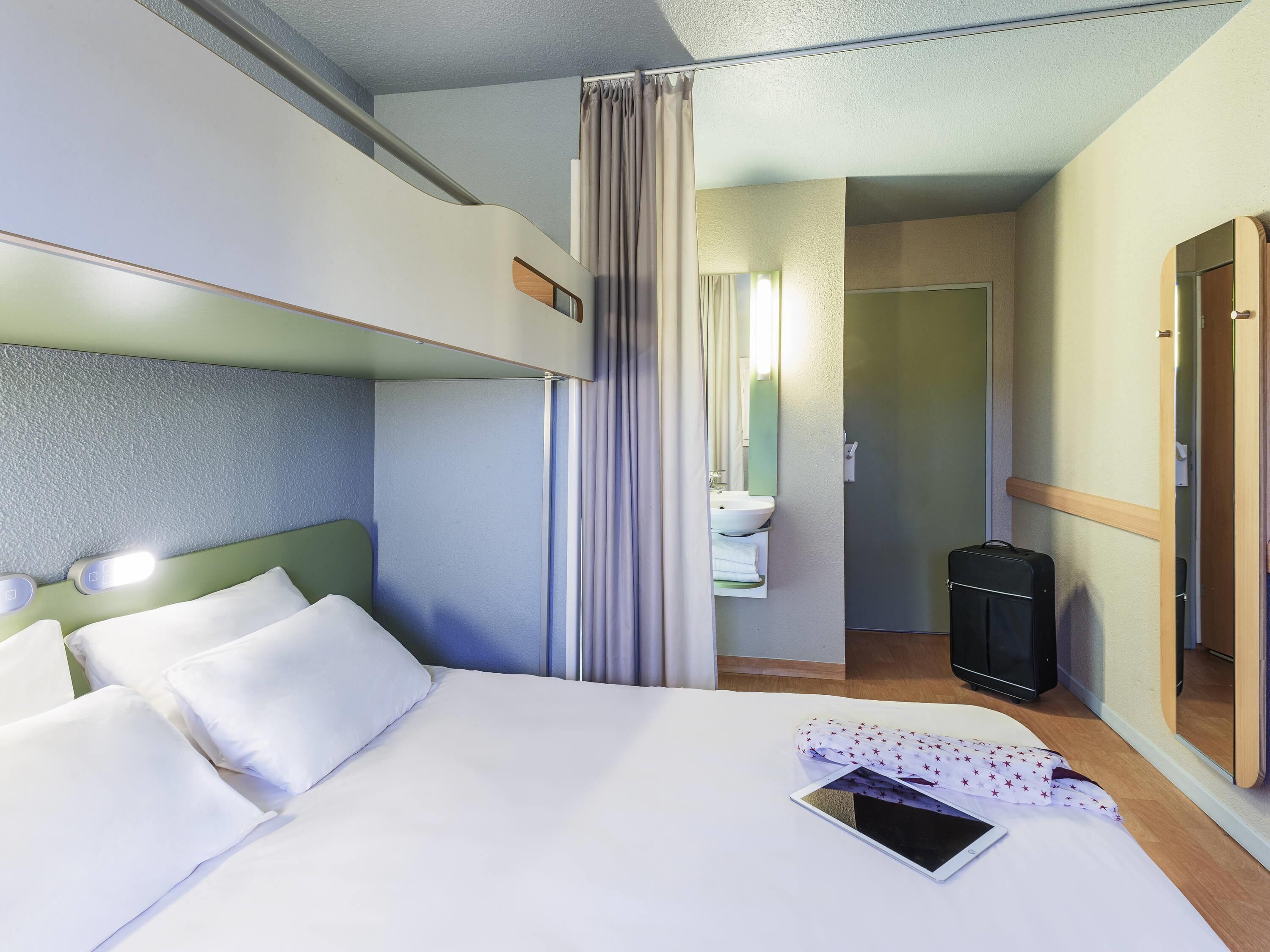 Una habitación de hotel con literas, una mesita de noche con una tableta, un baño con lavabo y espejo, y una maleta en el suelo.