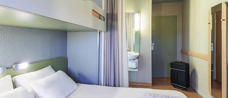 Una habitación de hotel con literas, una mesita de noche con una tableta, un baño con lavabo y espejo, y una maleta en el suelo.