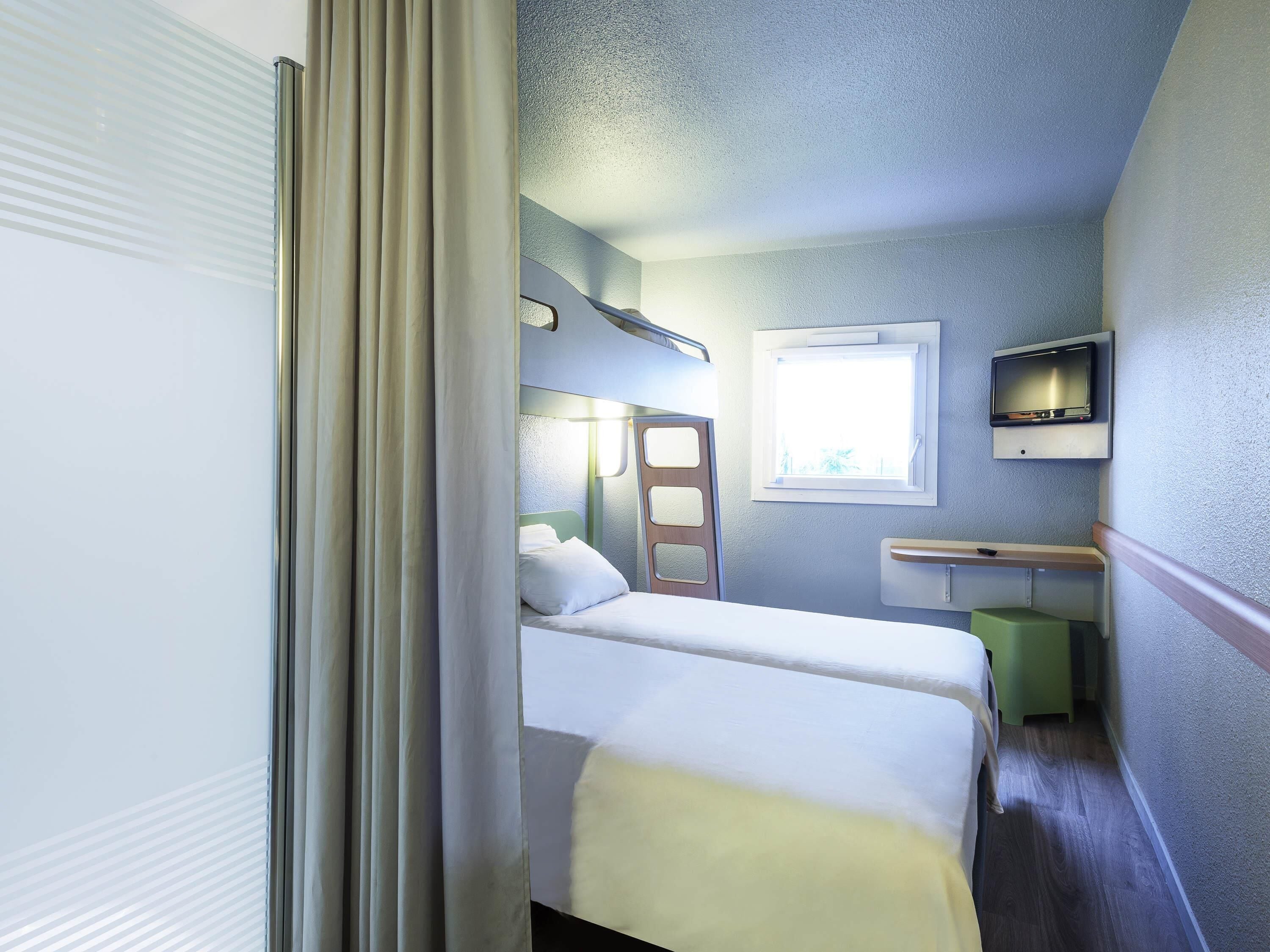 A compact hotel room with a bed, a small desk, a chair, a window, and a TV.