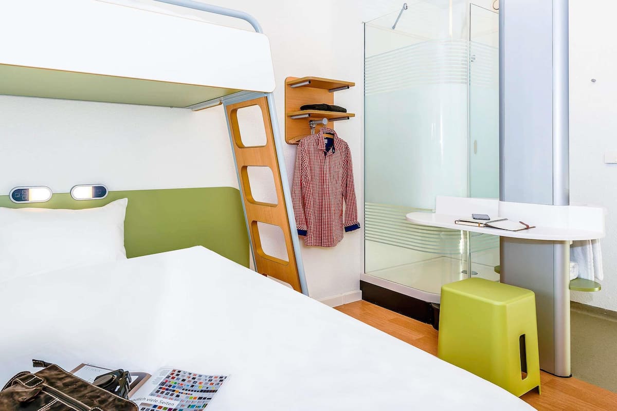 Une chambre d’hôtel avec un lit superposé, une petite salle de bain dotée d’une douche en verre, une table de chevet avec une lampe et un tabouret vert.