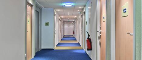 Pasillo de hotel con puertas numeradas 102 y 103, alfombra azul y señalización de salida de emergencia.