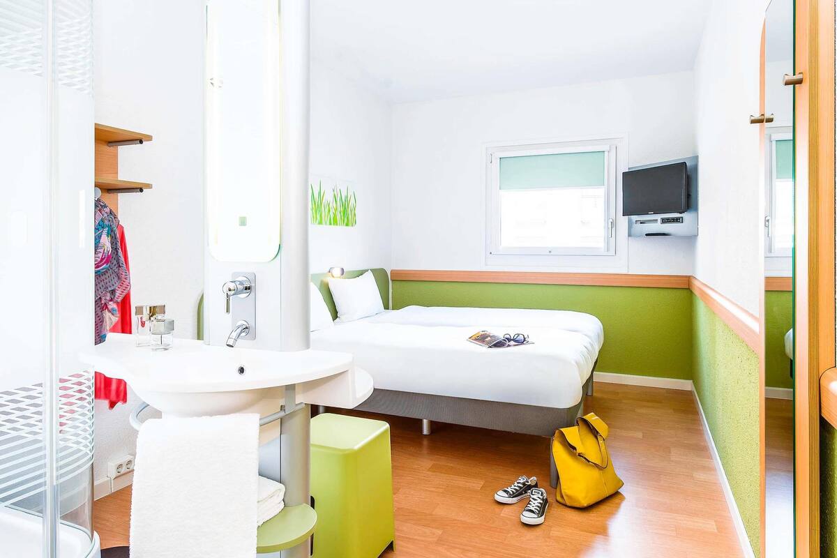 Une chambre d’hôtel avec un lit, un téléviseur fixé au mur, un petit bureau avec une chaise et une fenêtre.