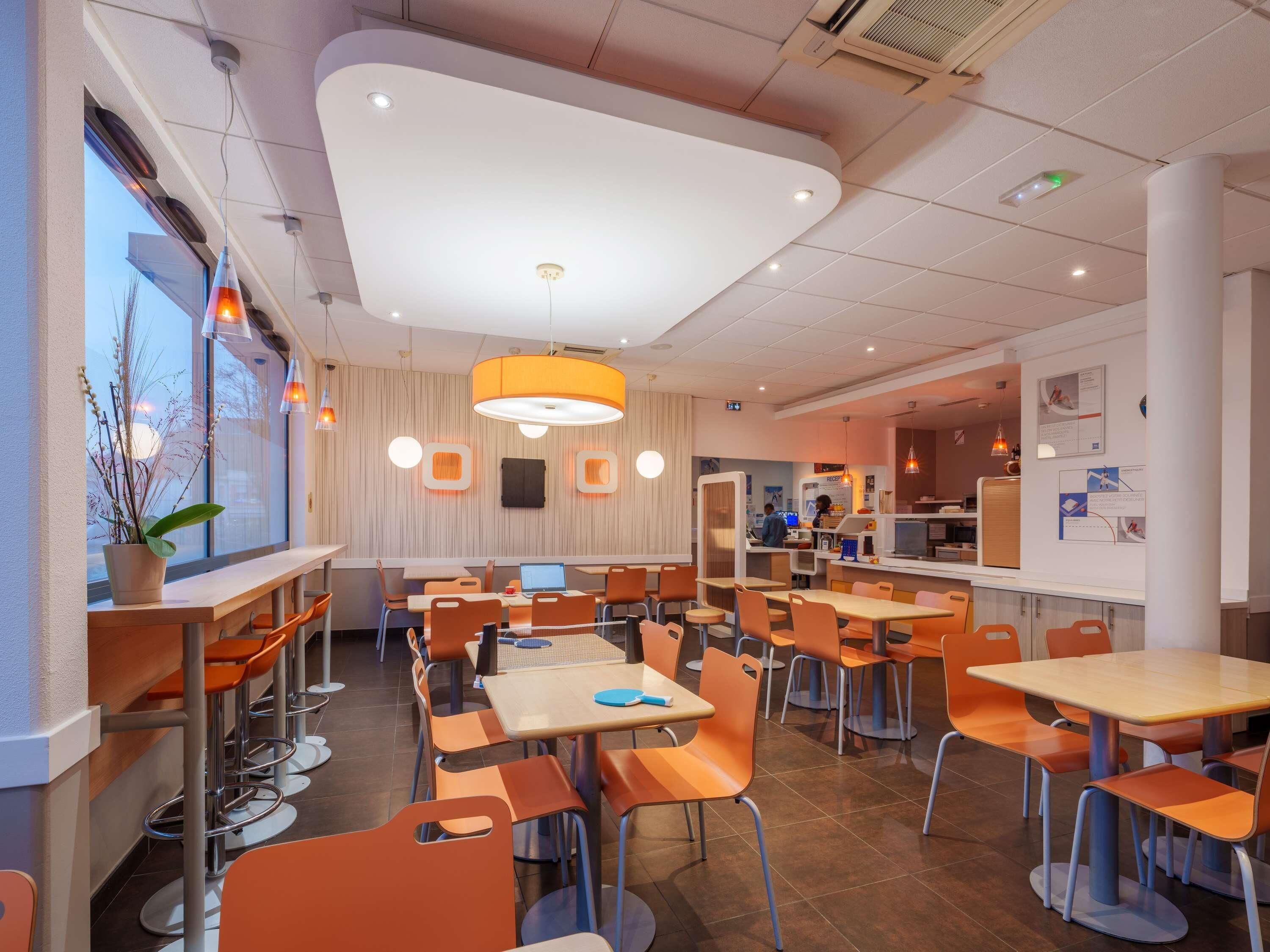 A modern restaurant with high tables and orange chairs, a large window with plants, and a bright ceiling.