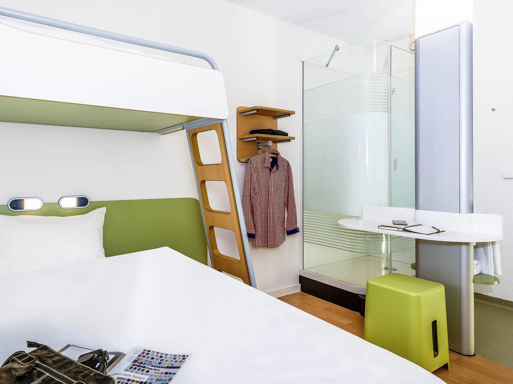A hotel room with a bunk bed, a small bathroom with a glass shower, a bedside table with a lamp, and a green stool.