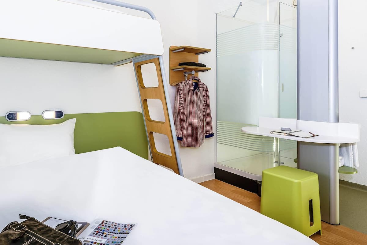 A hotel room with a bunk bed, a small bathroom with a glass shower, a bedside table with a lamp, and a green stool.