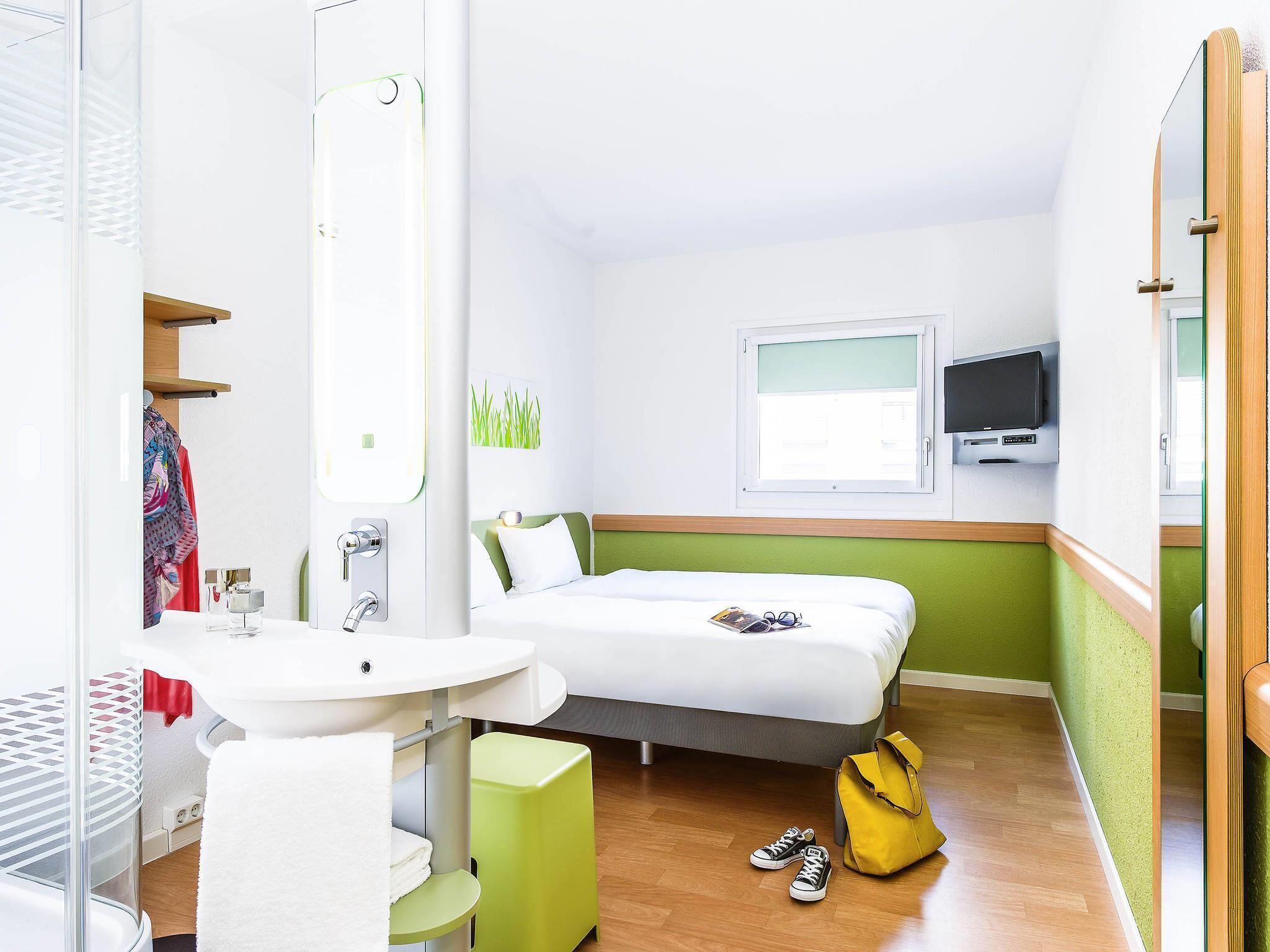A hotel room with a bed, a TV, a window, a bathroom with a sink and mirror, and a green stool.