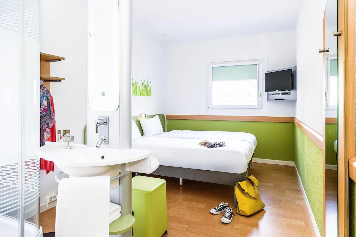 A hotel room with a bed, a TV, a window, a bathroom with a sink and mirror, and a green stool.