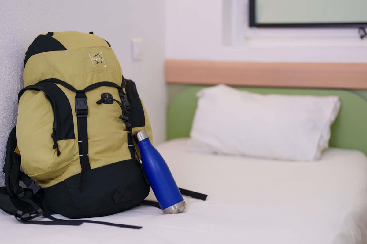 Un lit avec un oreiller blanc et un sac à dos jaune sur lequel est posée une bouteille d’eau bleue.