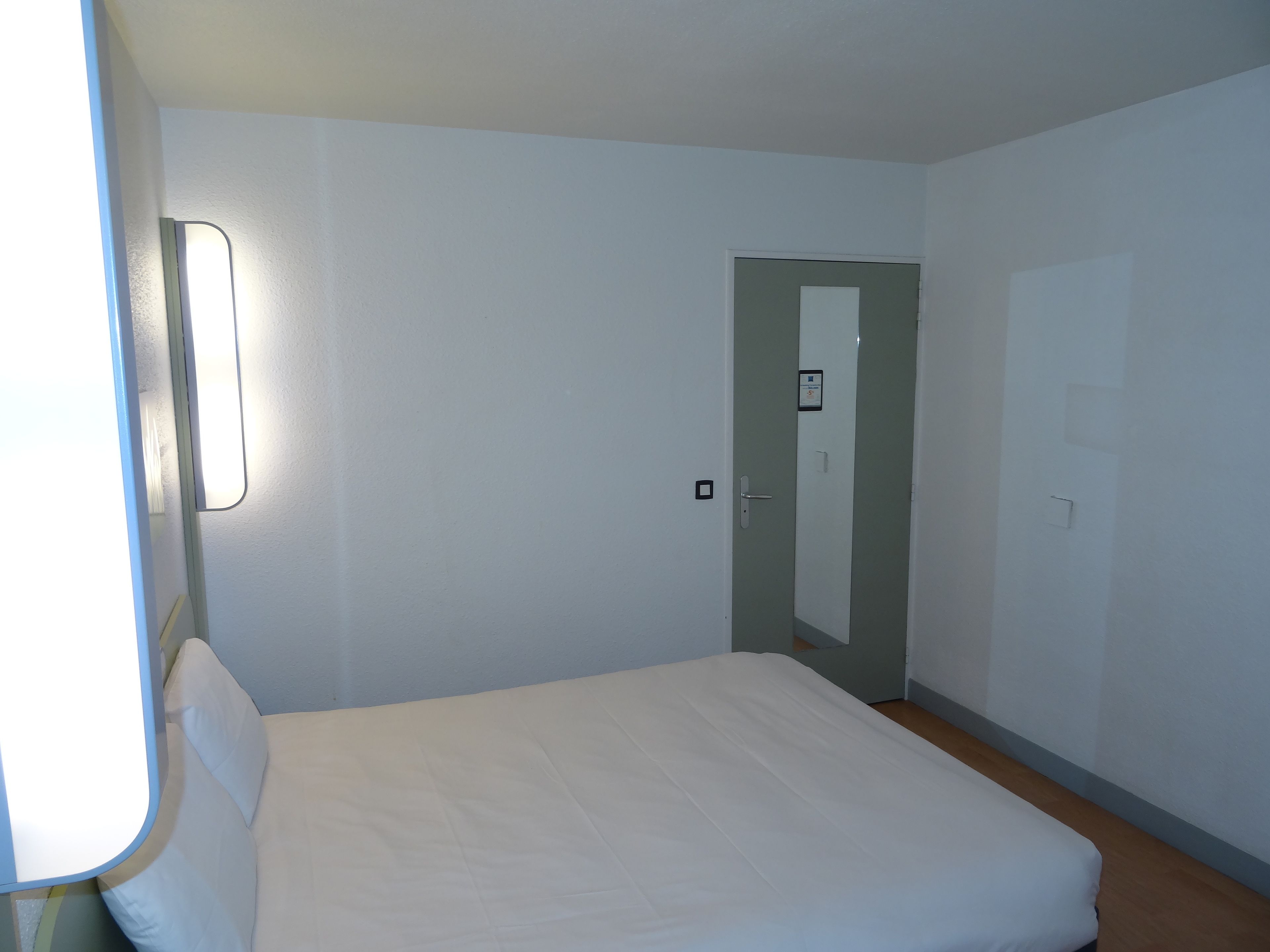 A simple, unadorned room with a bed, a door, and a light switch on the wall.