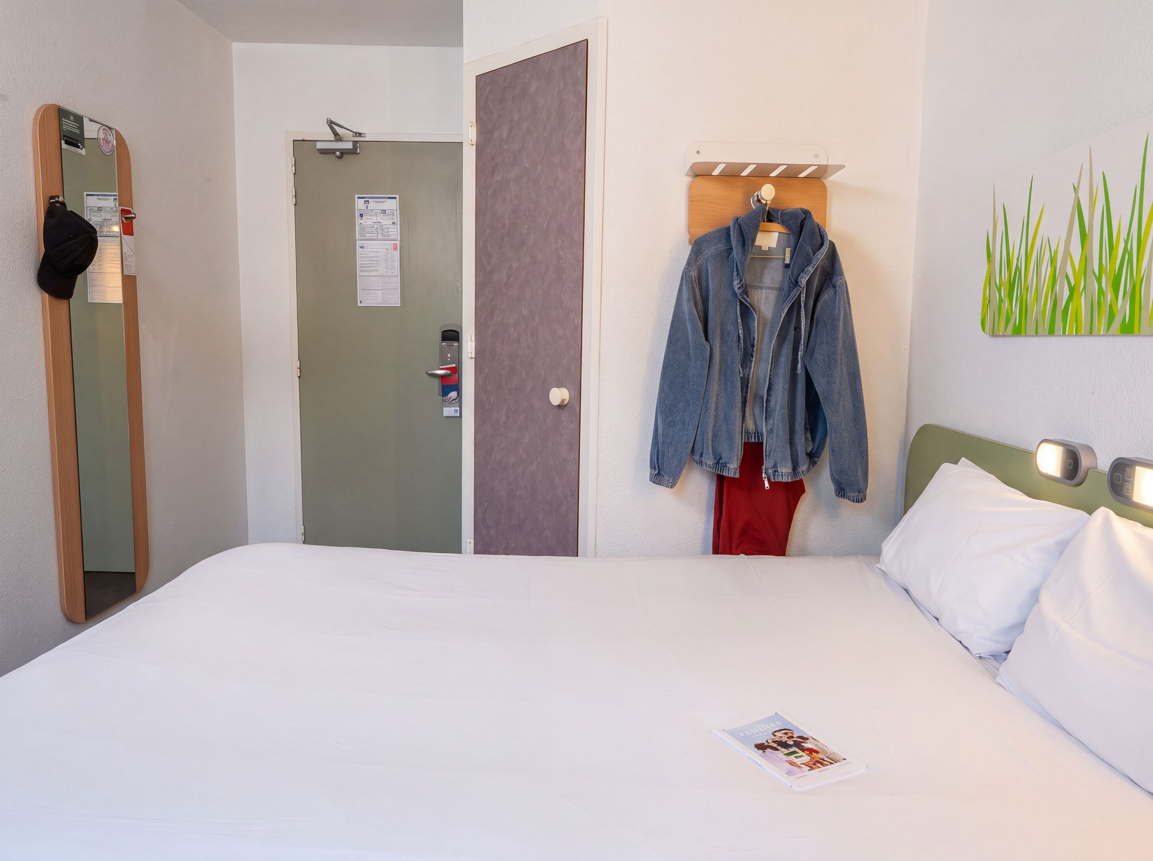 Habitación de hotel con cama, perchero, una puerta y una lámpara fijada a la pared.