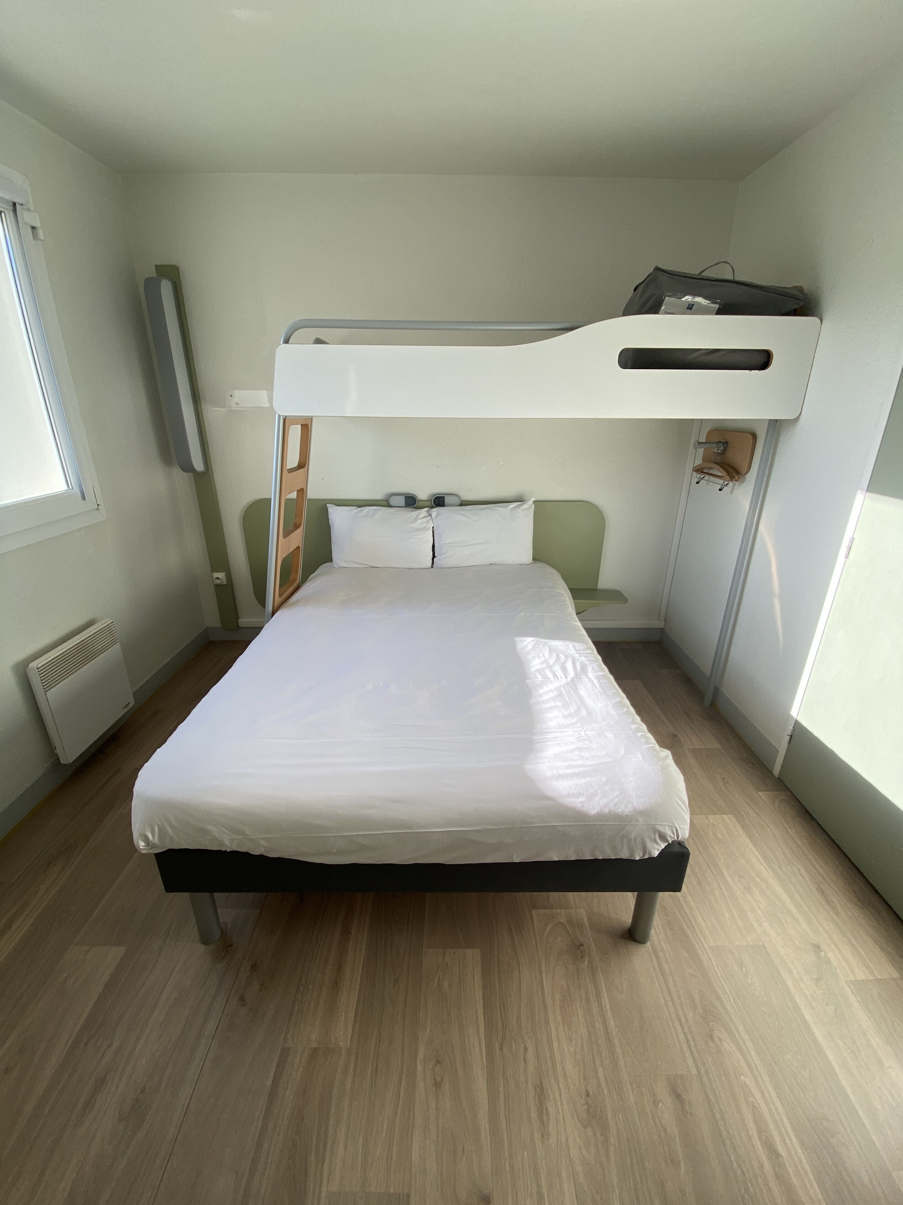 A bunk bed room with a white mattress, a wooden floor, and a window on the left.