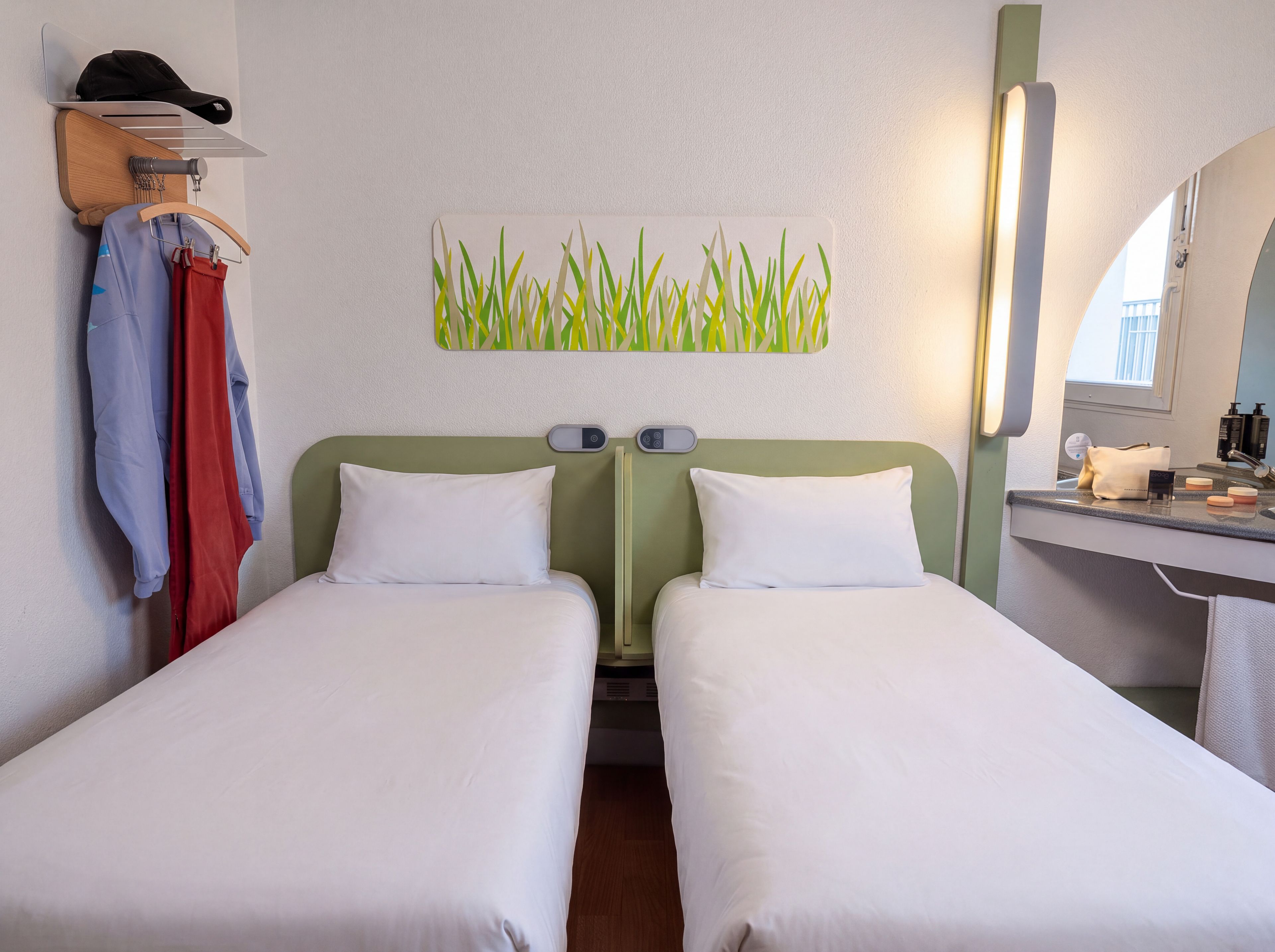 A hotel room with two single beds, a wall-mounted towel rack, a mirror, and a small bedside table with toiletries.
