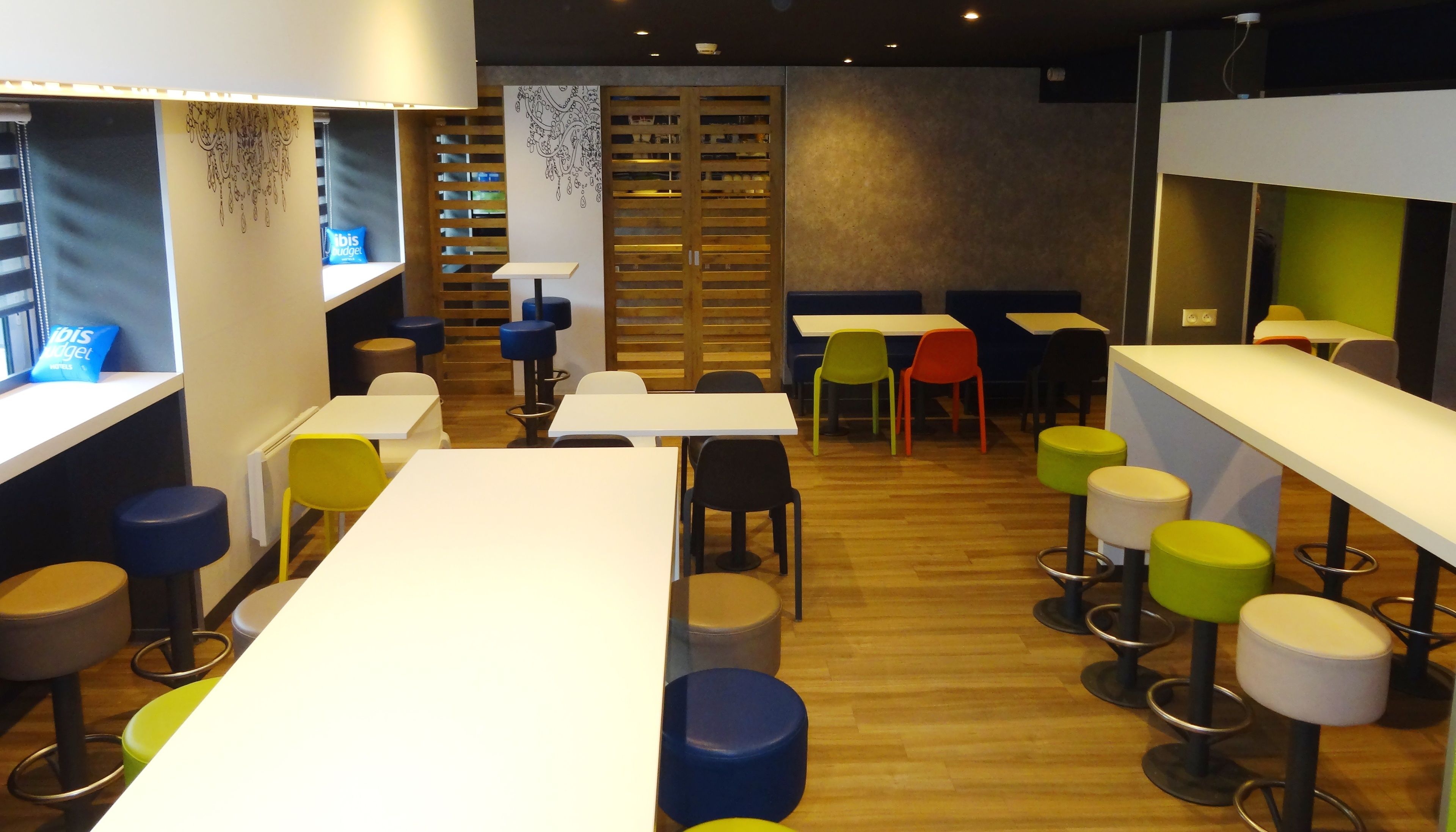 A modern cafe with white tables and colorful stools, wooden slatted partitions, and a patterned wall design.