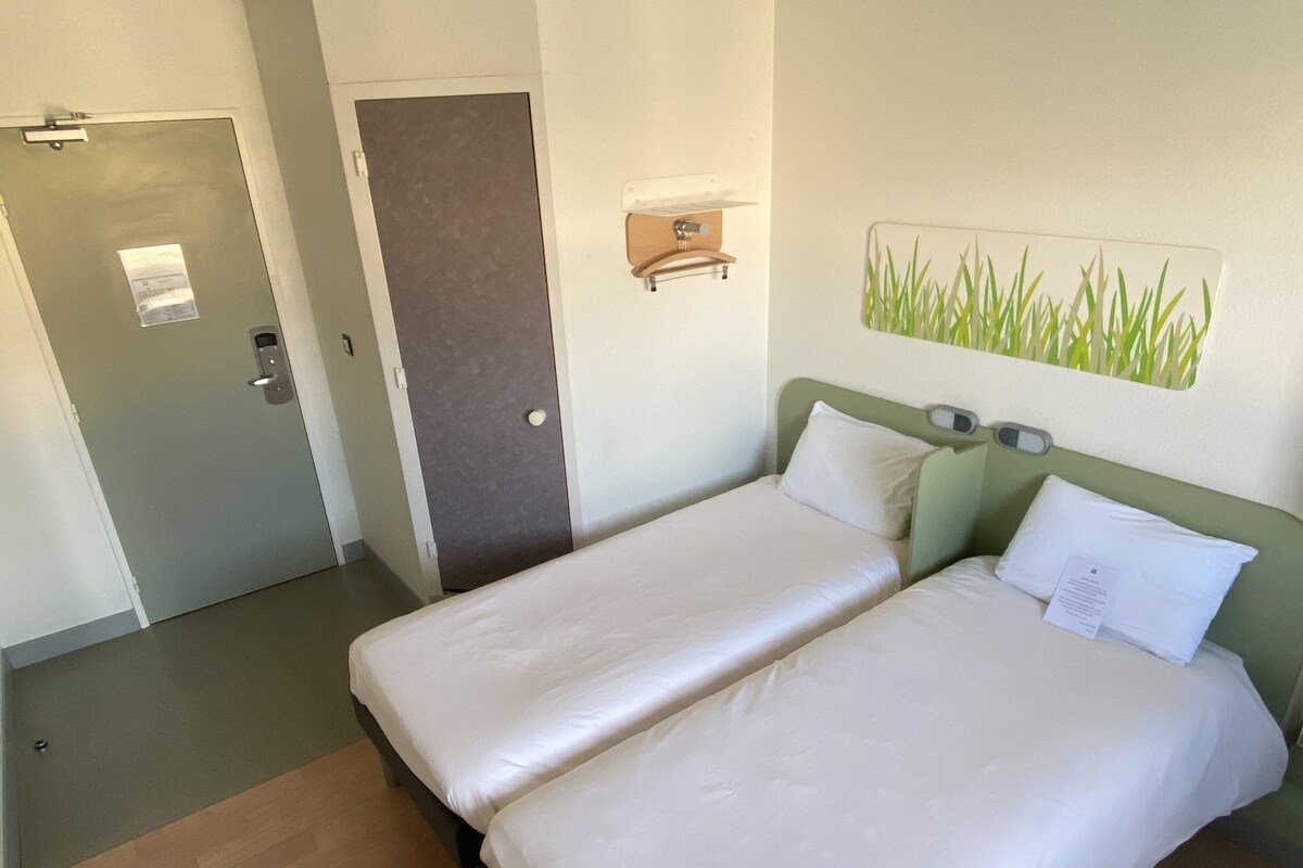 A hotel room with two single beds, a wooden floor, a door with a handle, and a wall-mounted light fixture.