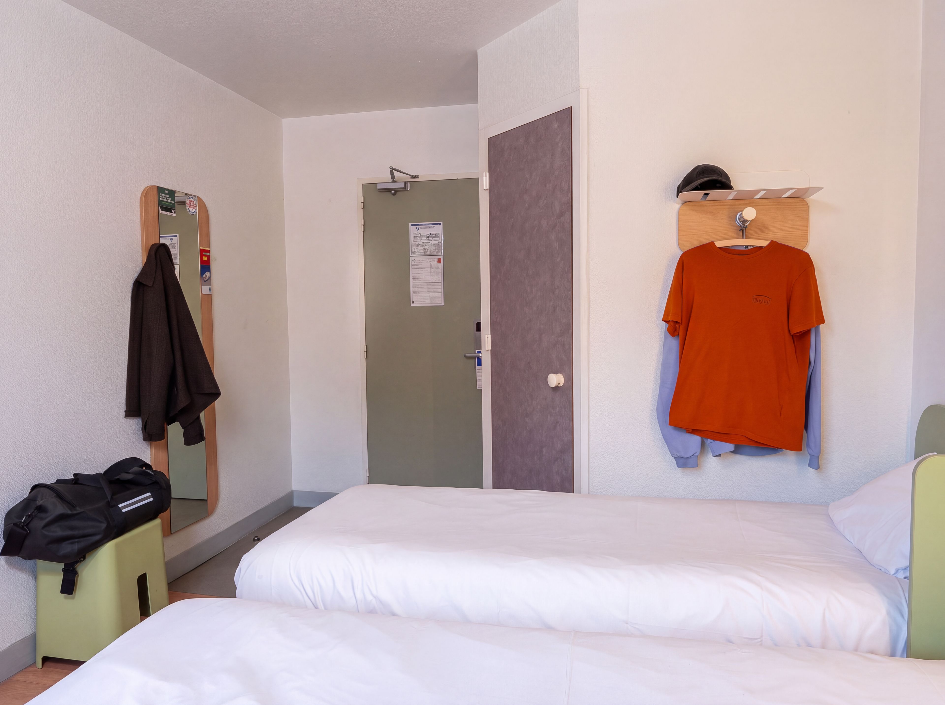 A hotel room with two beds, a mirror, a door, and a coat rack with a jacket and a shirt.