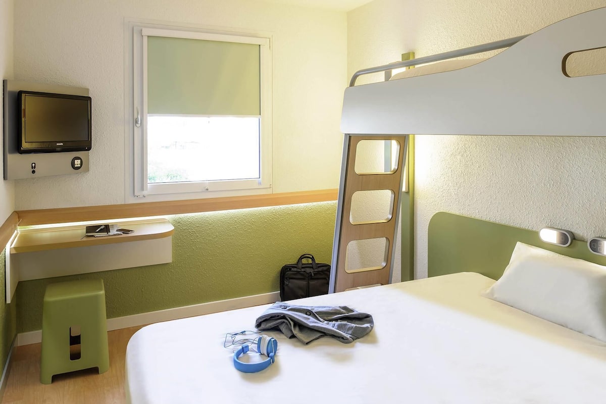 A hotel room with a bunk bed, a desk with a chair, a television, and a window.