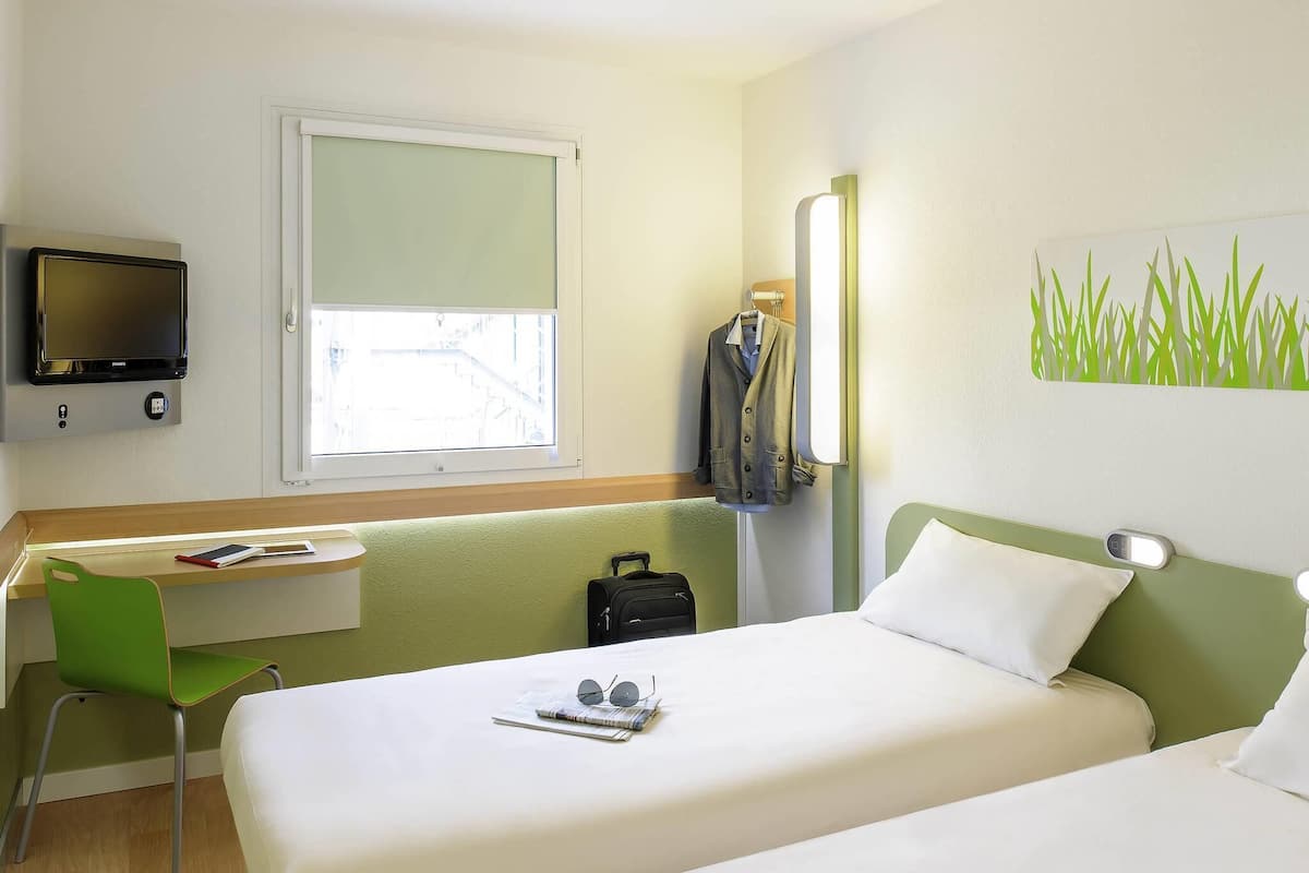Habitación de hotel con dos camas, un escritorio, una silla, un televisor y ventana con vistas a edificios.