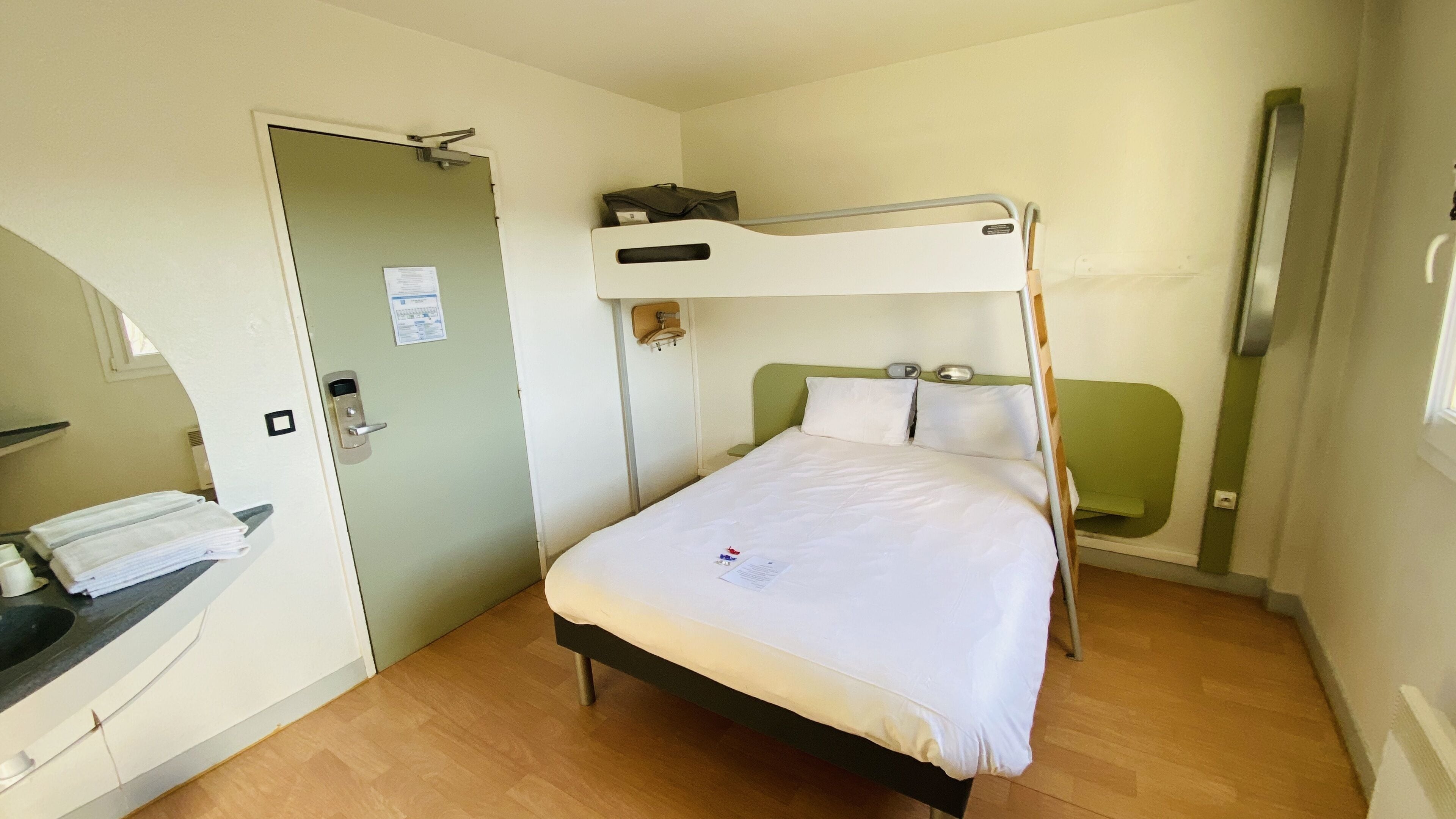 Habitación individual con una cama nido en alto, una mesita de noche, un escritorio pequeño y una puerta con cerradura.