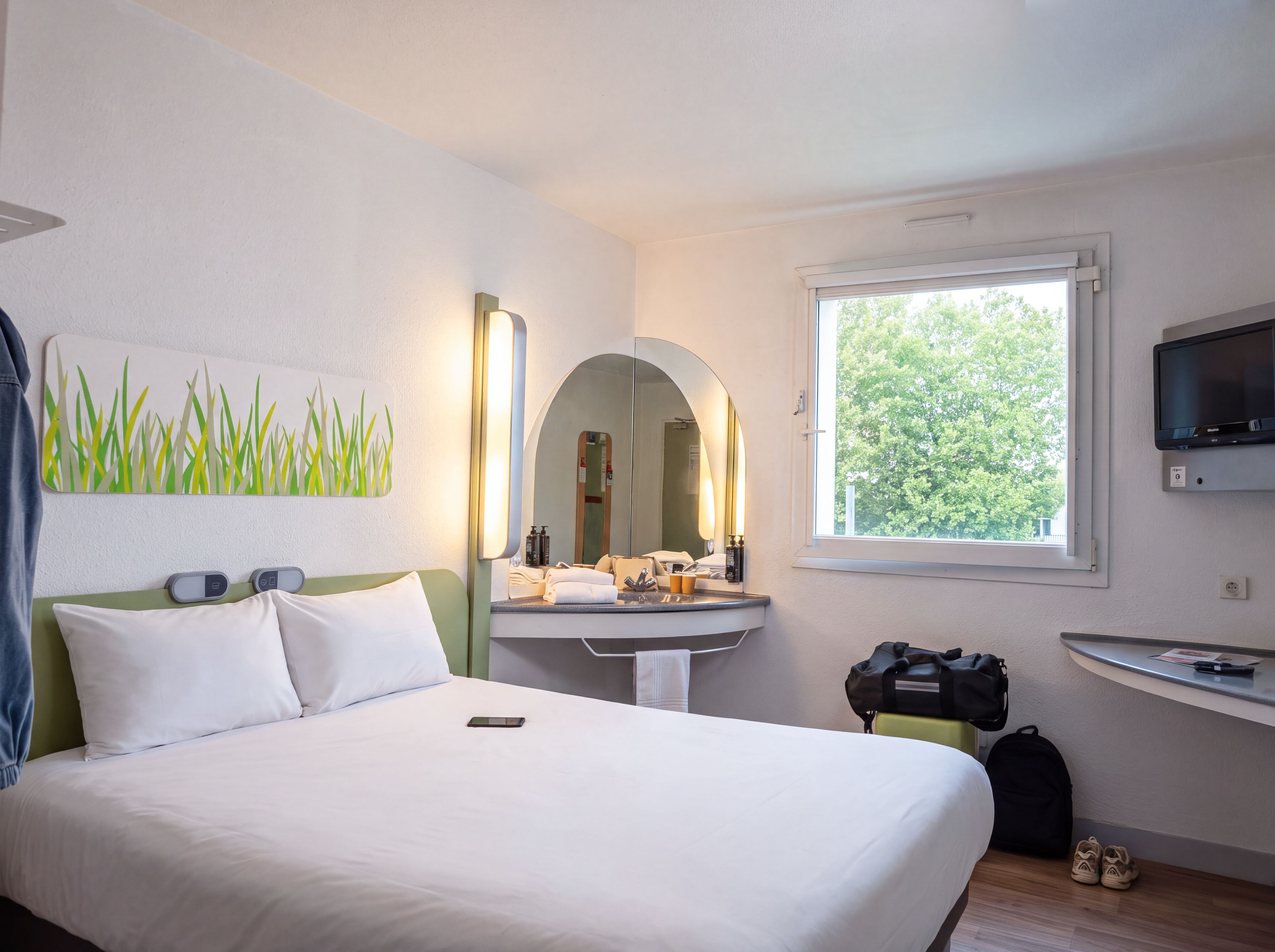 Habitación de hotel con cama, tocador con espejo, ventana con vistas a árboles y televisor montado en la pared.