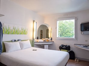 A hotel room with a bed, a vanity with a mirror, a window with a view of trees, and a TV mounted on the wall. - ibis budget Caen Centre Gare (Caen)