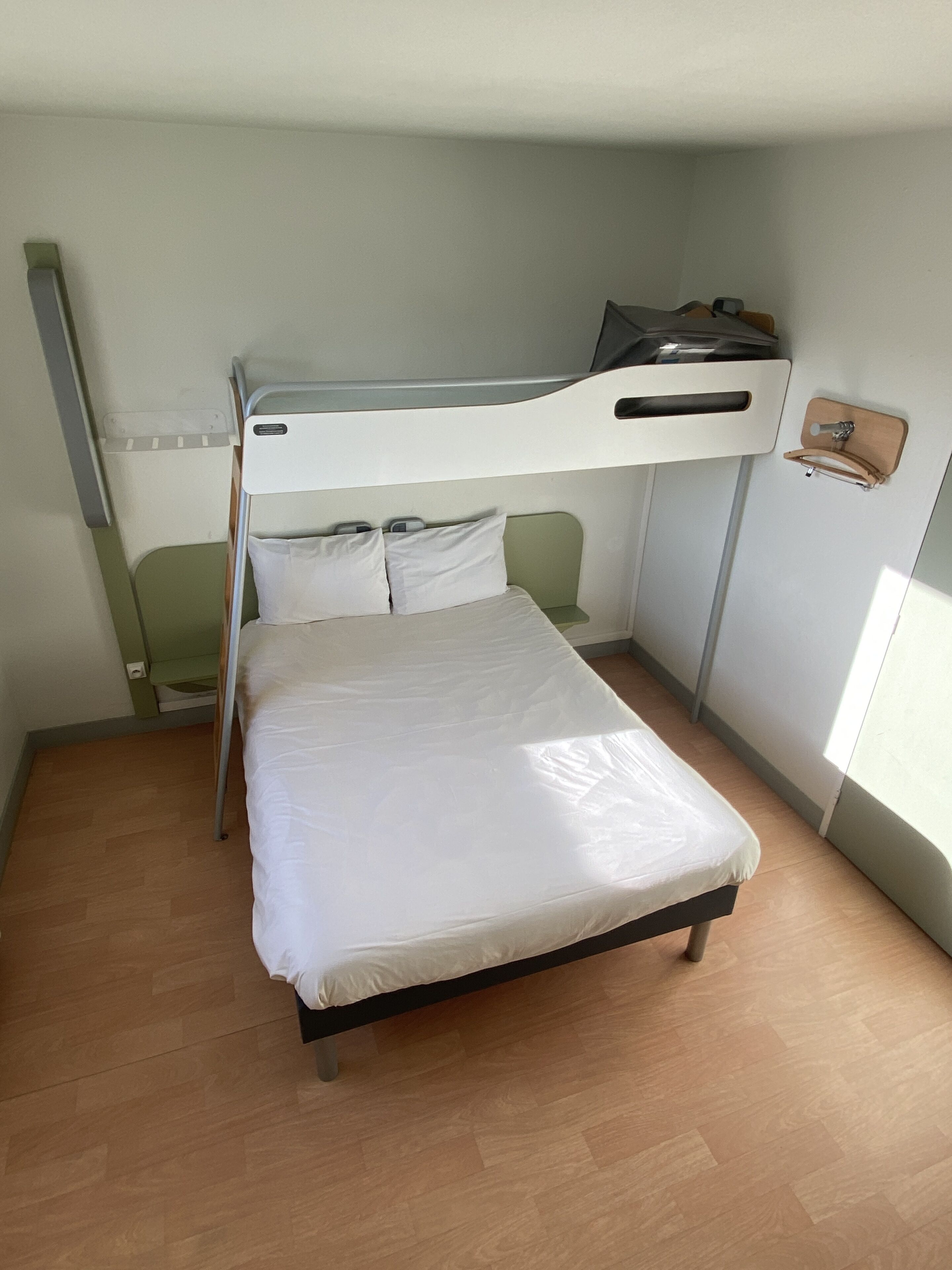 A small, single-bed room with a bunk bed above, a wooden floor, and a white wall with a small shelf.