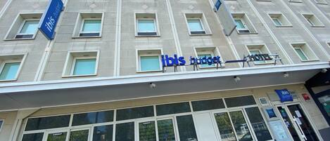 Fachada de hotel con múltiples ventanales, un llamativo letrero de 'ibis budget' y un cartel de 'CANAL+' que indica una ubicación cercana.
